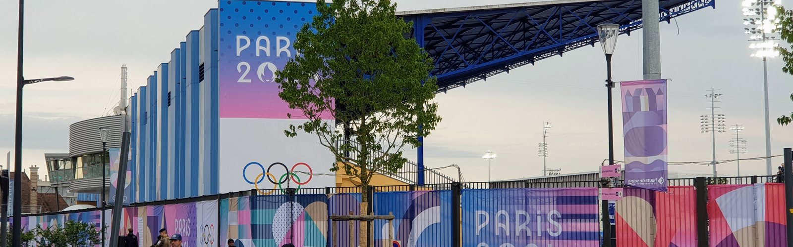 Paris 2024 hoardings and the Olympic rings along a stadium fence